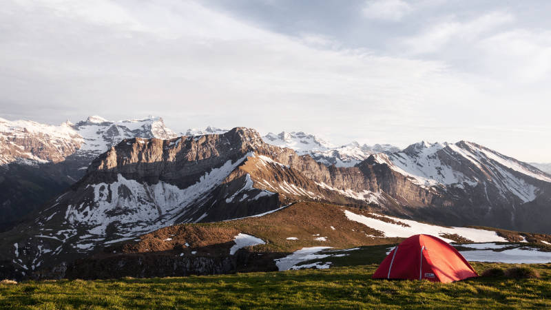 
Ein rotes Zelt auf einem grasbewachsenen Hügel mit Bergen im Hintergrund.
