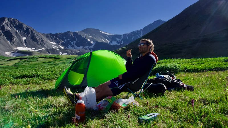 Ein Mann unternimmt einen Solo-Campingausflug und baut sein grünes Zelt inmitten einer malerischen Berglandschaft auf und isst sein mitgebrachtes Essen.